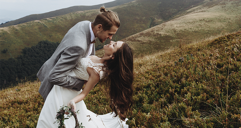 Romantic wedding session of a bride and groom in the mountains - bride with garland and groom in grey jacket in a creative pose during outdoor wedding photography. An example of natural and beautiful wedding photos of the bride and groom.