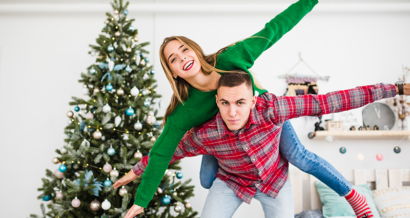 A boy having a girl up on his shoulders. A Christmas tree and a bed in the background.