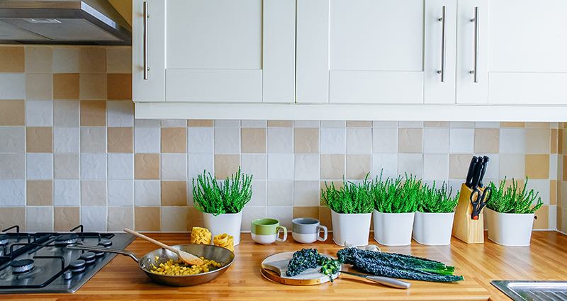 A close-up on a bright kitchen counter top with herbs in white flower pots and colourful vegetables on it. White cupboards over the counter top.