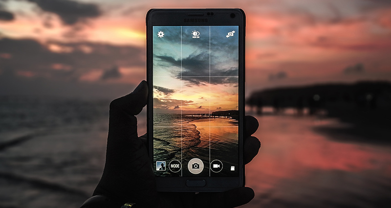 Le zoom sur une main tenant une smartphone, sur l’écran le coucher du soleil visible au fond.