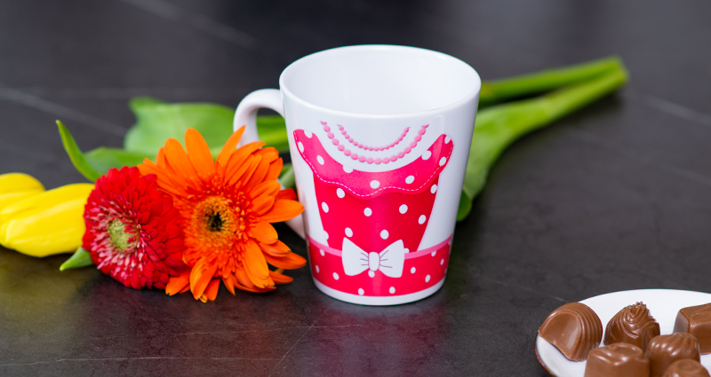 Un mug latte avec le motif de robe rose à pois, à côté les fleurs de printemps et des chocolats sur une assiette.