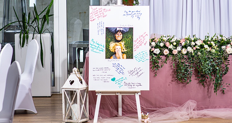 Didelė drobė su įsimylėjusia pora viduryje, aplink vestuvių svečių sveikinimai parašyti spalvingais žymėkliais. Šalia baltas žibintas, o fone stalas, puoštas rožinės spalvos medžiaga ir gėlėmis