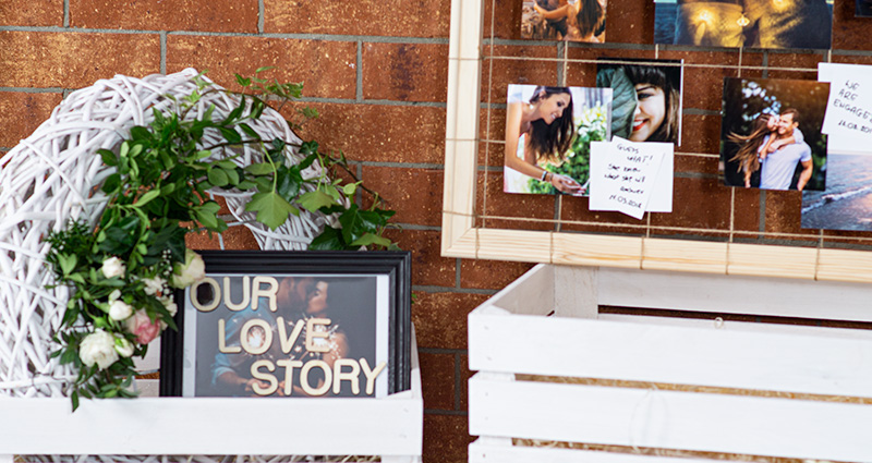       Priartinimas ant užrašo „Our Love Story“ juodame rėme, dekoruoto gėlėmis, baltoje medinėje dėžutėje, šalia medinio DIY rėmo fragmentas su jaunavedžių nuotraukomis iš skirtingų gyvenimo etapų su komentarais. Plytų siena fone.
