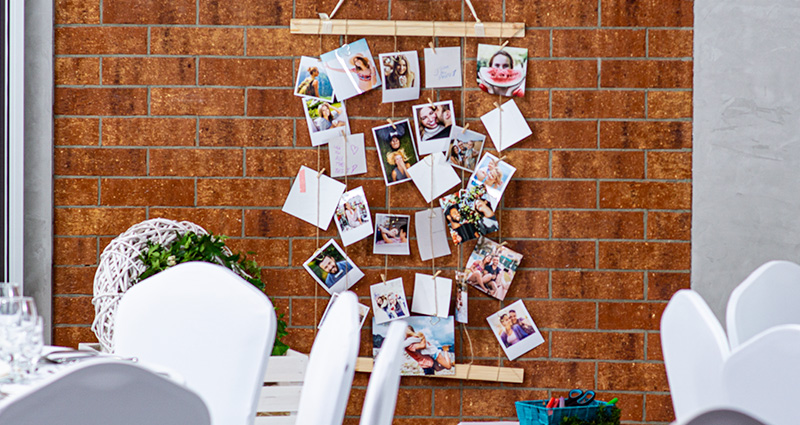 Insta-Fotos mit Hochzeitsgästen, die an einer Juteschnur auf Holzleisten an einer Ziegelwand befestigt sind. Im Vordergrund sind die Hochzeitstische in weißer Farbe dekoriert.