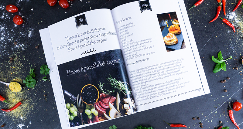 Un fotolibro aperto – un ricettario, con intorno alcuni peperoncini, dei pomodorini e delle spezie.