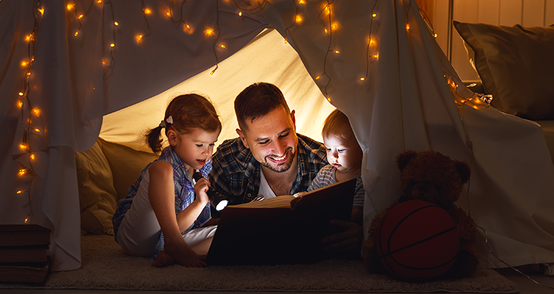 Un padre che legge una storia ai propri bambini dentro una tenda fatta con le lenzuola e illuminata da piccole lucine.