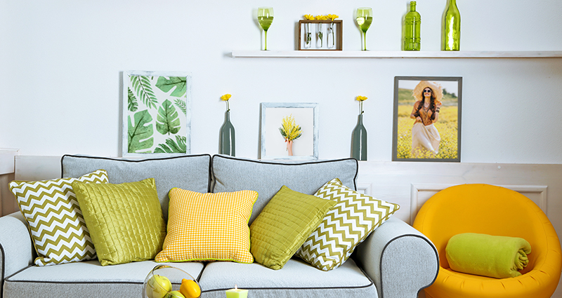Nuotrauka pilkos sofos su žaliomis ir geltonomis pagalvėlėmis įvairiais modeliais, šalia geltonas fotelis. Ant stalo vaisiai, o už sofos 3 pavasariniai paveikslai ir įvairūs aksesuarai.