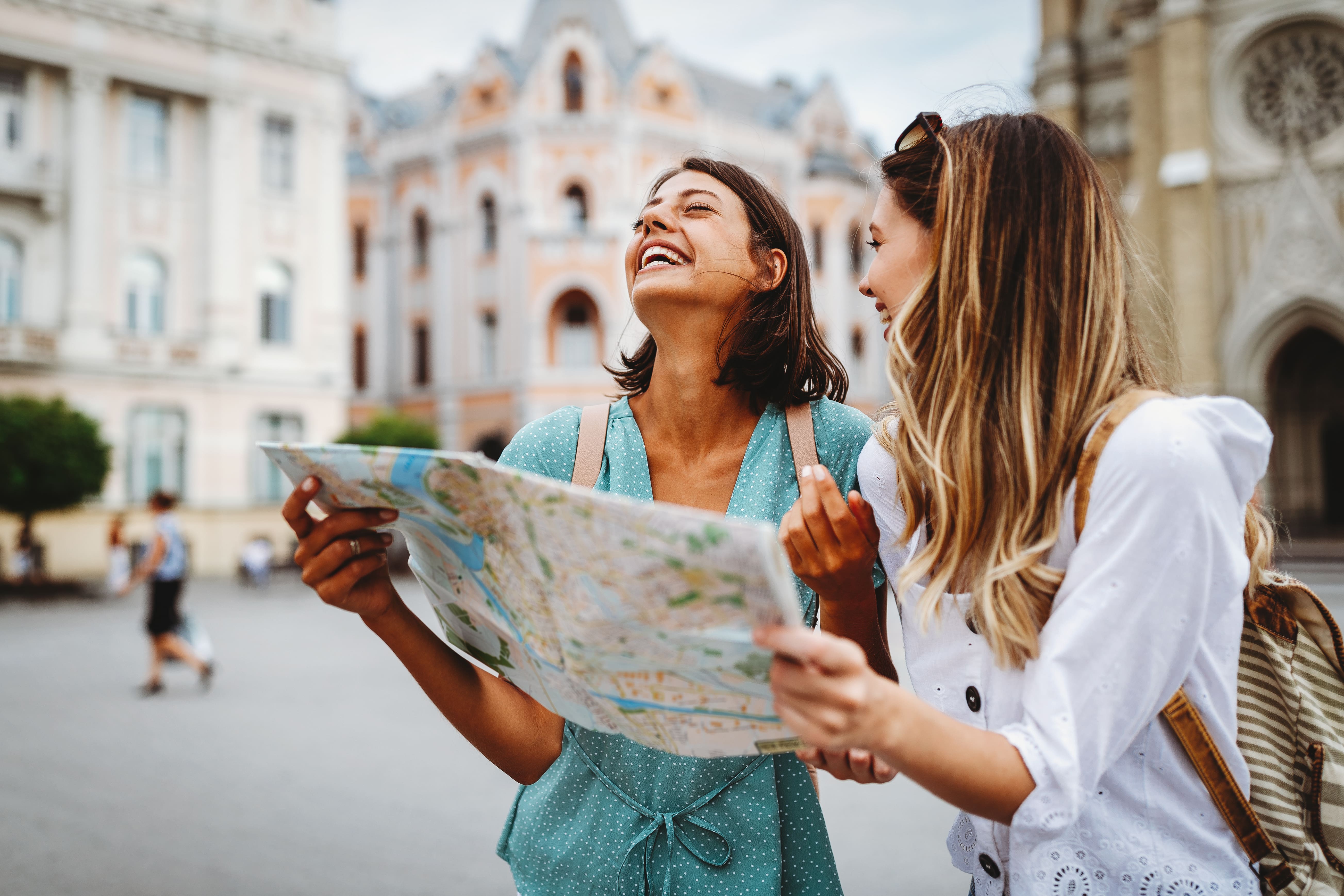 Zwei junge Frauen in einem historischen Stadtzentrum halten eine Karte. Statt für das Foto zu posieren, lachen sie und sehen entspannt aus, als hätten sie einen unserer Reisefotografie Tipps zu Herzen genommen.