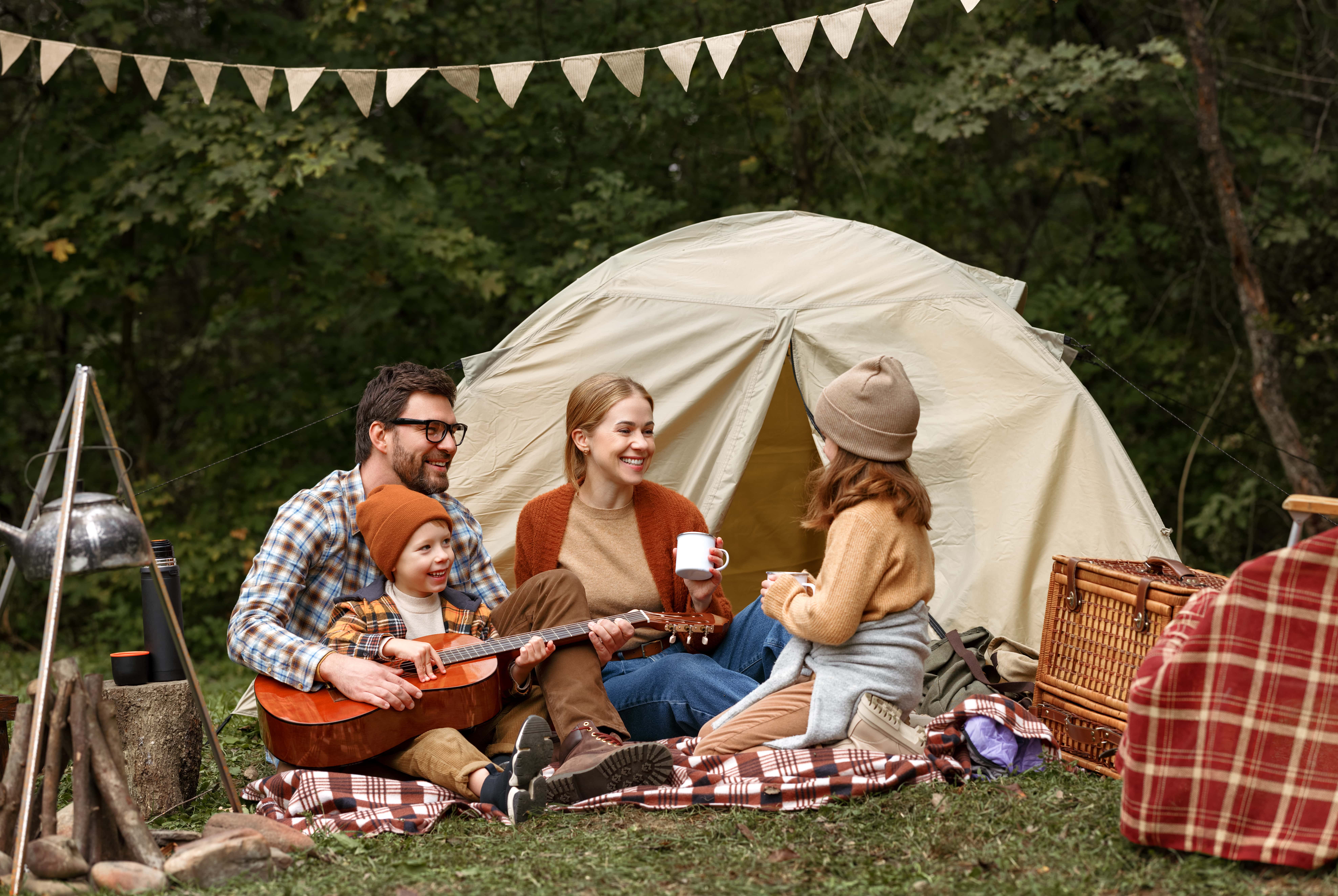 Eine Familie sitzt vor einem weißen Zelt vor den grünen Bäumen. Die Mama und ihre Tochter trinken etwas Warmes, während der Papa mit dem Sohn, der eine Gitarre auf dem Schoß hat, spielt. Eine ideale Szenerie, um viele schöne Urlaubsbilder zu machen.