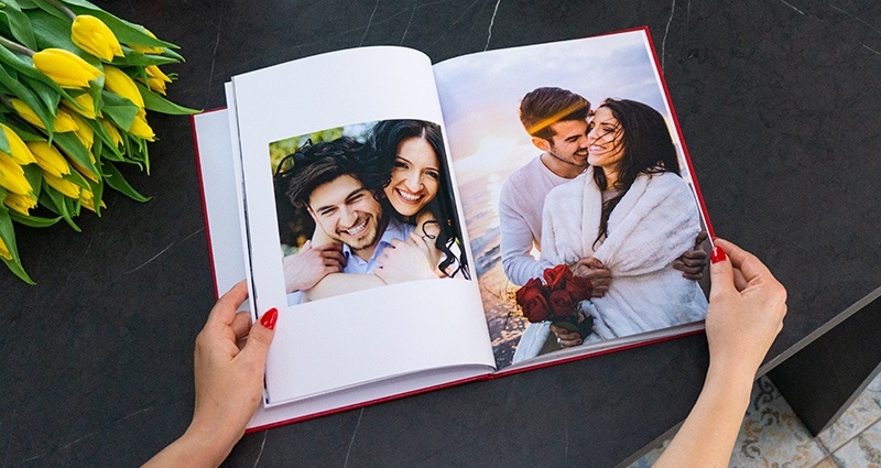 Inzoomen op de handen van een vrouw, die een open fotoboek met afbeeldingen van een paar vasthoudt; naast twee fotomokken, een ander cadeau dat wordt gebonden met een rode strik en een boeket gele tulpen.