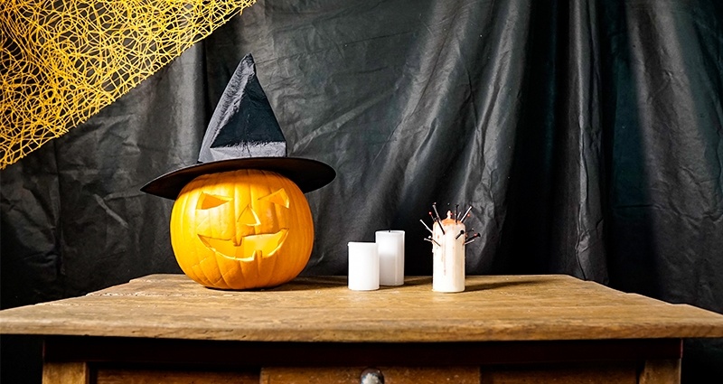 Halloween pumpkin in a witch hat put on the old brown table next to 3 candles, the black curtain in the background and the orange net in the corner