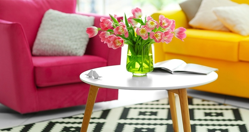 Foto de una mesa con una jarra con tulipanes rosas, al lado una alfombra blanca y negra, en el fondo un sillón de púrpura y una sófa amarilla.