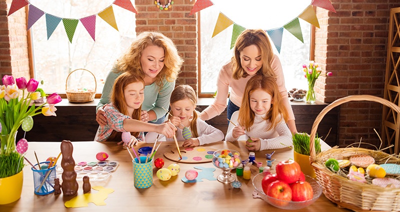 Drei Mädchen und zwei Frauen an einem Tisch voller Eier, Pralinen, Farben und Tulpen.