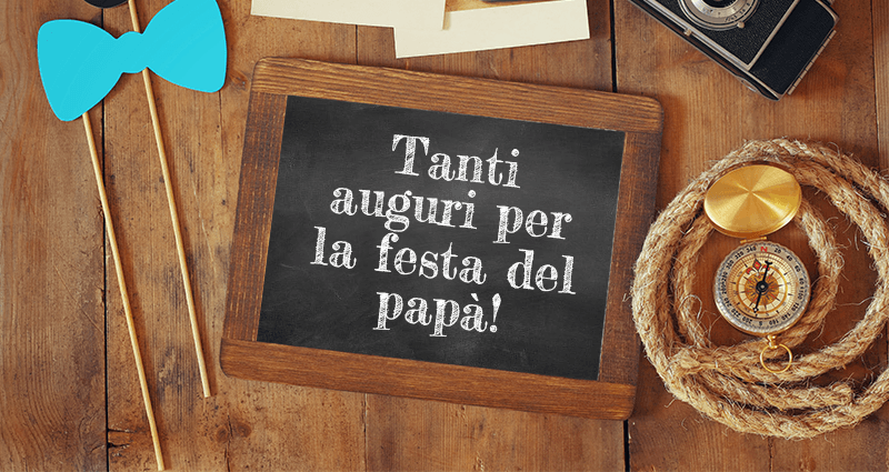 Una piccola lavagna in legno con la scritta Buona Festa del Papà con accanto una serie di oggetti appoggiati su una scrivania in legno una macchina fotografica, una corda, alcune stampe retro.